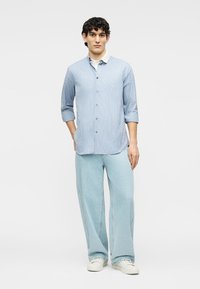 Νεαρός άνδρας που στέκεται με τα χέρια στις τσέπες, φορά ένα ανοιχτό μπλε ριγέ πουκάμισο με κουμπιά, άνετο ανοιχτό μπλε παντελόνι και λευκά αθλητικά παπούτσια.