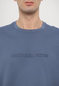 Kék pamut környakú póló, amelyen kiemelkedő betűkkel van írva: "MICHAEL KORS" az elején. Simított textúra és laza kialakítás.