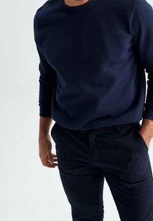 Homme portant un pull à manches longues bleu foncé et un pantalon noir, debout avec une main dans la poche devant un fond clair uni.