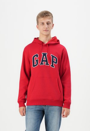 Red hoodie with "GAP" logo in navy, featuring a front pocket and drawstring hood. Made of soft fabric; fitted design. Blue denim jeans worn.