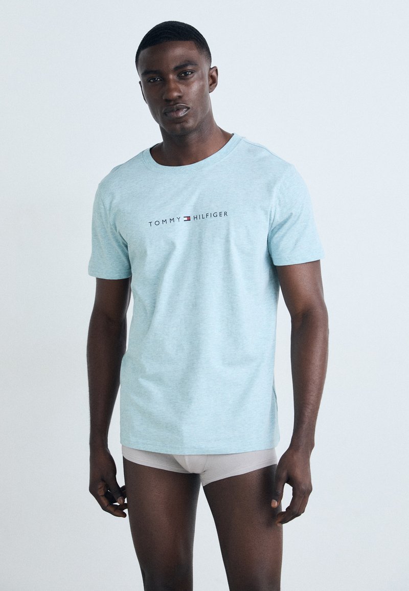 Šviesiai mėlynas medvilninis marškinėliai su apvaliu kaklu ir "Tommy Hilfiger" logotipu ant krūtinės. Modelis po jais dėvi smėlio spalvos šortus.
