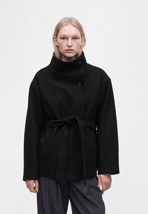 Young person with straight blonde hair wearing a belted black coat with high collar and dark loose-fitting pants against a plain background.