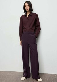 Bordeaux-rode blouse met knopen, gecombineerd met donkerpaarse brede broek met witte zijkanten, aangevuld met lichte schoenen.