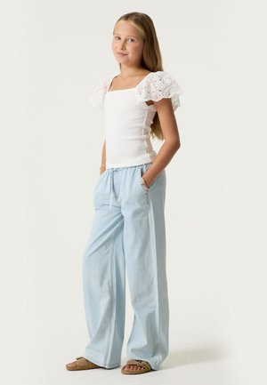 Niña joven de pie con las manos en los bolsillos, vistiendo una blusa blanca de manga abullonada, pantalones anchos de pierna en azul claro y sandalias marrones sobre un fondo liso.