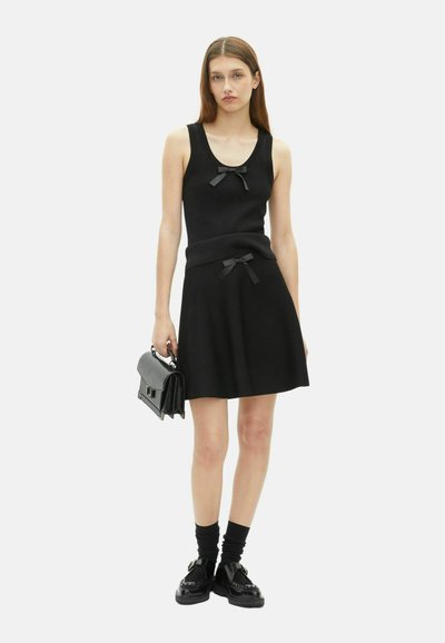 Robe noire avec un haut ajusté et une jupe évasée, ornée de deux nœuds noirs décoratifs. Associée à un sac à main noir et des chaussettes épaisses.