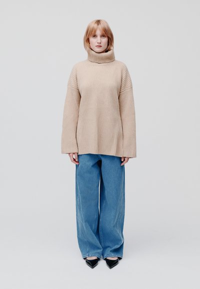 Beige geribbelde coltrui met verlaagde schouders, gecombineerd met lichtblauwe wijde jeans en puntige zwarte hakken.