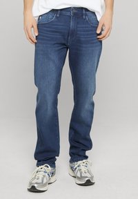 Jean denim bleu foncé avec une coupe ajustée, cinq poches et passants pour ceinture. Légère décoloration le long des cuisses et coupe droite des jambes.