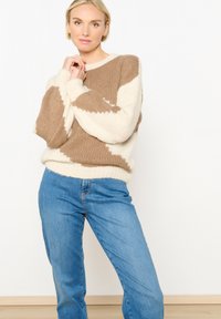 Pletena svetrovka z zasnovo, ki vključuje rjave in bele bold zigzag vzorce, okroglim izrezom in rebrastimi manšetami, kombinirana s svetlo modrimi jeans hlačami.