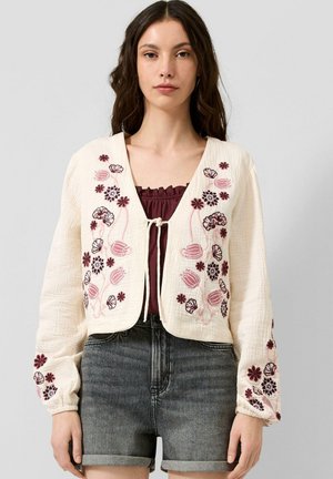Vrouw draagt een crèmekleurig vest met roze en bordeauxrode bloemenborduursel over een bordeaux top en donkere omgeslagen spijkershorts.