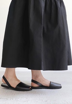 Czarne sandały na obcasie typu slingback z otwartym palcem, wykonane z gładkiej skóry, na płaskiej podeszwie, o minimalistycznym designie, w zestawieniu z czarną, zwiewną spódnicą.