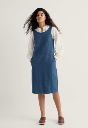 Hosszú sötét hajú nő térdig érő, ujjatlan farmerruhát visel fehér, hosszú ujjú blúz fölött, barna bebújós cipővel.