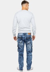 Felpa grigia e jeans cargo blu con finitura invecchiata, numerose tasche e cerniere. Taglio casual e rilassato. Sneakers con suola in gomma.