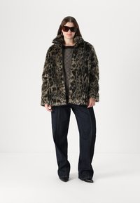 Plaščenka iz umetnega krzna z leopardjim vzorcem in visokim ovratnikom, nosena čez črn pulover z vzorcem, v kombinaciji s temnimi kavbojkami in čevlji s peto.