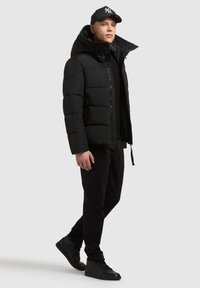 Chaqueta de plumas negra con cuello alto, cierre de cremallera y diseño acolchado. Usada sobre una camiseta negra, combinada con pantalones negros y zapatillas.