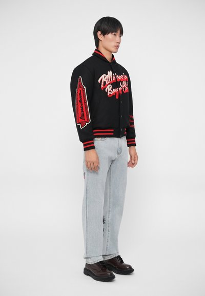 Jaqueta varsity preta com bordados vermelhos e punhos listrados, combinada com jeans azul claro e sapatos castanhos escuros com atacadores.