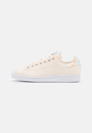 Beige lage sneaker met witte zool, vetersluiting, geperforeerde zijstrepen en bloemaccent op het hielstuk.
