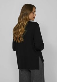 Pull en tricot noir à manches longues, avec une texture côtelée et des fentes latérales au bas, associé à un pantalon large gris. Vue de dos.