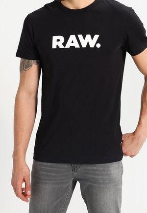 Mężczyzna w czarnej koszulce z wyraźnym białym napisem "RAW. ", w szarych dżinsach i widocznym tatuażu na górnej lewej ręce.