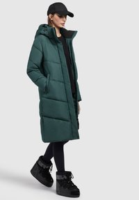 Cămașă lungă verde căptușită cu glugă, cu fermoar în față și buzunare laterale, purtată peste un top negru și leggings negri, asortată cu cizme negre până la glezne.