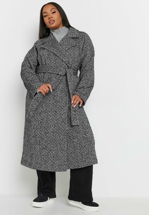 Schwarz-weißer Übermantel mit Fischgrätmuster, einem taillierten Gürtel, oversized Ärmeln und Reverskragen; getragen über einem grauen Rollkragenpullover.