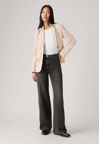 Blazer beige ajusté sur un t-shirt blanc, associé à un jean noir taille haute et ample. Textures lisses et détails de design minimalistes.