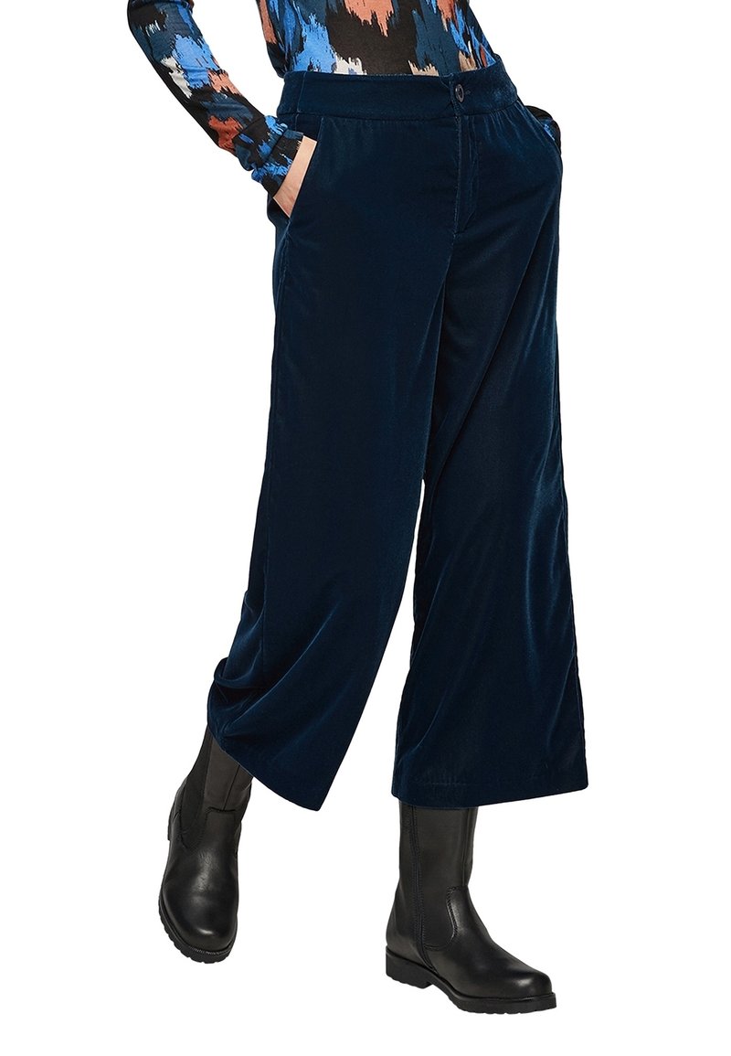 Personne portant un pantalon large en velours bleu marine, des bottines en cuir noires, et un haut à manches longues à motif abstrait, les mains dans les poches.