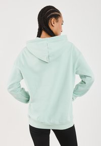 Sieviete ar pītām matām, ģērbusies gaiši piparmētru zaļā kapucē un melnās bikses, stāv ar rokām uz gurniem, skatoties prom pret baltu fona.