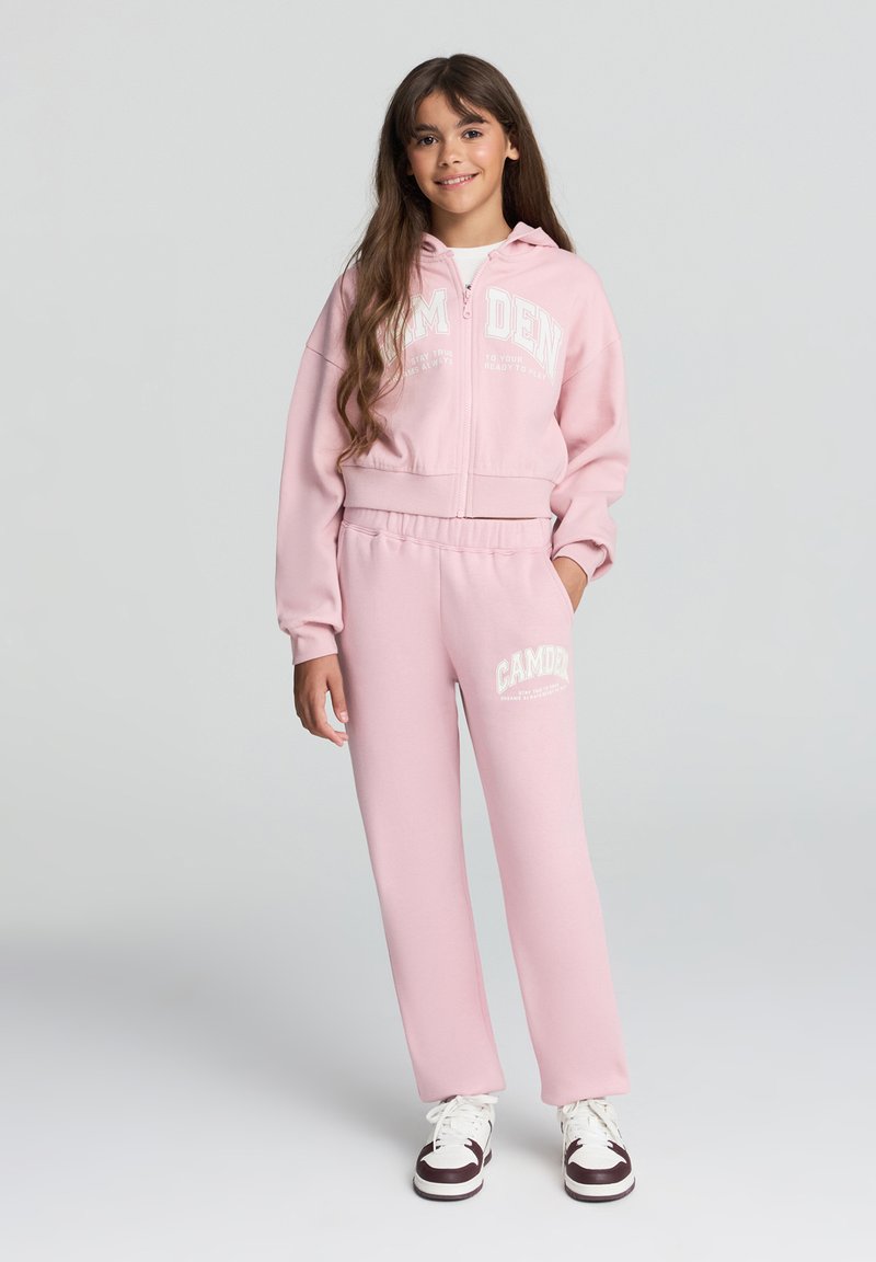 Felpa rosa con grafica bianca "CAMDEN", chiusura con zip, e pantaloni della tuta rosa abbinati con dettaglio logo. Accenti bianchi sulle calzature.