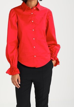 Vrouw draagt een felrode blouse met knopen, pofmouwen en gerimpelde manchetten, gecombineerd met zwarte strakke broek.