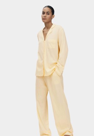 Vrouw staand, gekleed in een losse, lichtgele pyjamabovenkleding met knopen en borstzak, met bijpassende wijde broekspijpen, tegen een effen achtergrond.