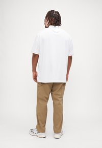 Chemise blanche à manches courtes avec une coupe décontractée, associée à un pantalon cargo beige et des baskets grises, vue de dos.
