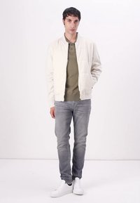 Leichte Bomberjacke aus Creme, gesteppter Design, mit gerippten Bündchen, kombiniert mit einem olivgrünen Pullover, grauen Jeans und weißen Sneakers.