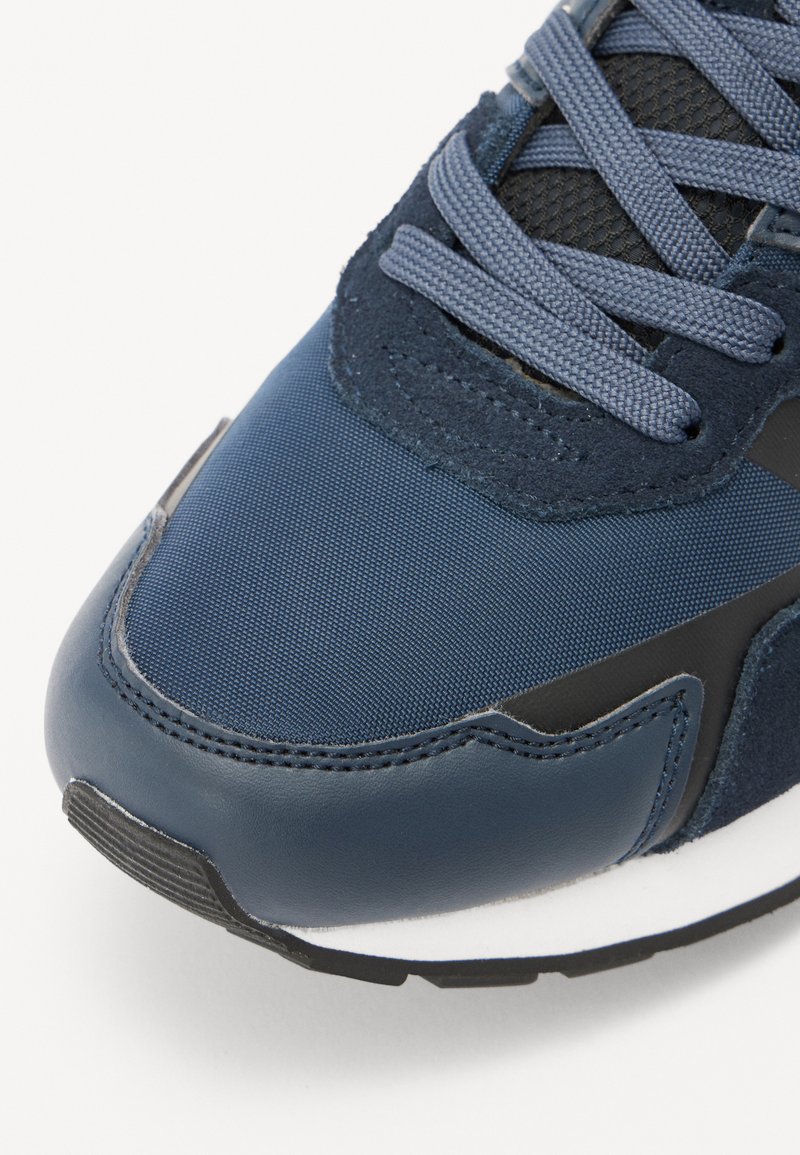 Scarpa da ginnastica blu con una combinazione di tessuto testurizzato e pelle, punta tonda, lacci spessi e suola bianca con accenti neri.