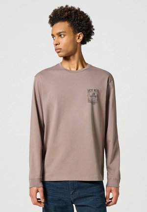 Joven con pelo rizado que lleva una camisa de manga larga color malva con el texto y gráfico "Hot Rod" en el pecho, combinada con jeans azules.