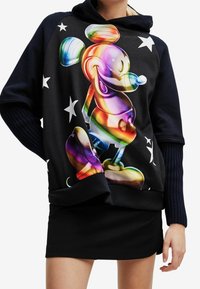 Sudadera negra con capucha que presenta un gráfico colorido de Mickey Mouse con acentos de arcoíris, rodeado de estrellas blancas. Detalle de mangas acanaladas visible.