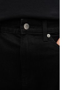 Κοντινό πλάνο της μέσης μαύρου τζιν με ασημένιο κουμπί και τρουκ, μερικώς εμφανιζόμενο χέρι στην τσέπη.