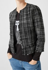 Mustavalkoinen ruudullinen lyhyt takki, jossa on pyöreä kaulus ja repaleiset reunat, kerrostettuna mustan yläosan kanssa, jossa on kissagrafiikka.