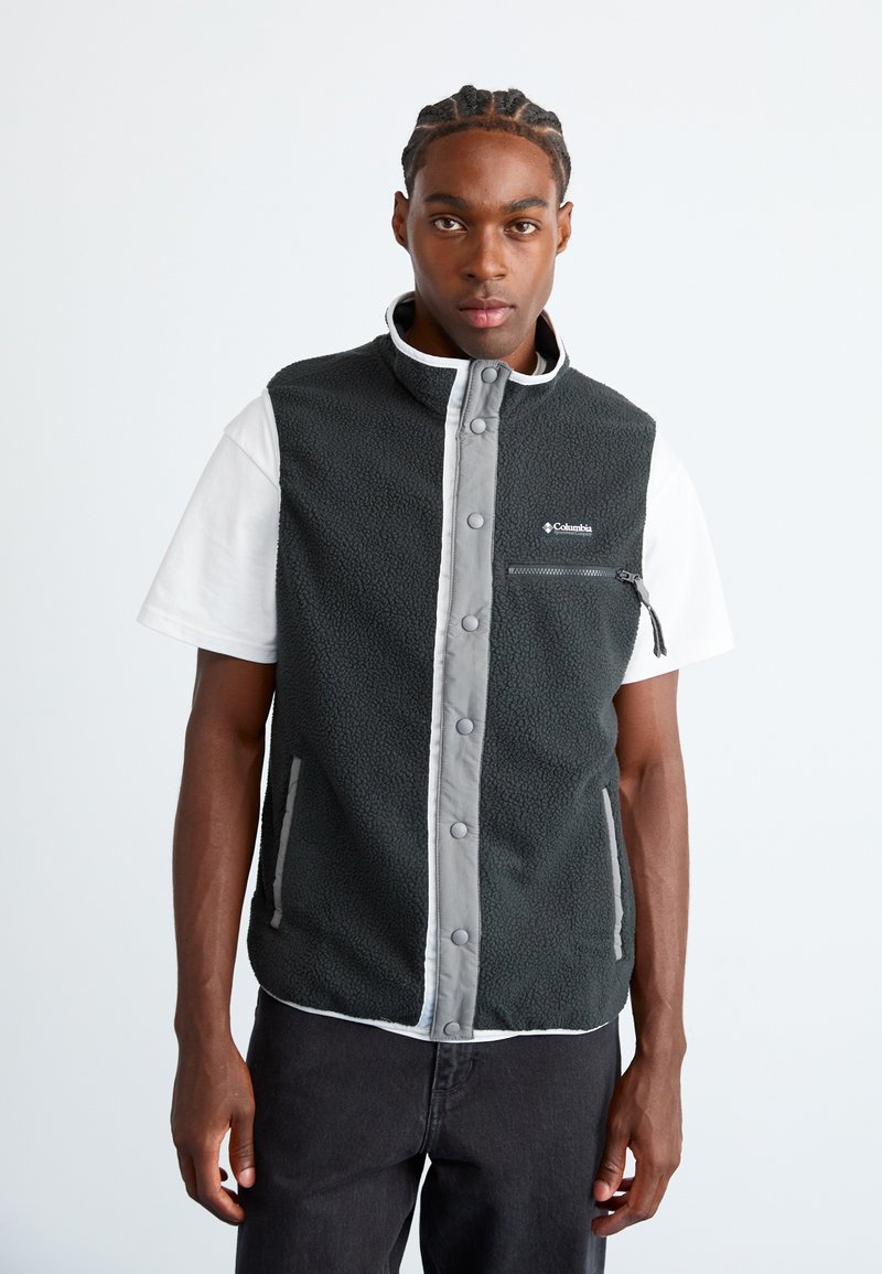 Ung mand med fletninger iført en sort fleece Columbia vest over en hvid t-shirt mod en ensfarvet lys baggrund.