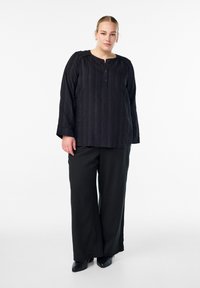 Crna bluzica dugih rukava s teksturiranim prugama i okruglim izrezom, s detaljem gumba sprijeda, u paru s slobodnim crnim hlačama i cipelama s oštrim vrhom.