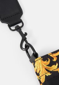 Crochet pivotant en métal noir reliant une sangle en cuir noir à un tissu à motif avec un design ornemental jaune.