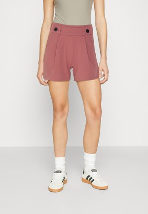 Femme portant un short taille haute rose poudré avec des boutons noirs, des chaussettes blanches et des baskets Adidas blanches à rayures noires.