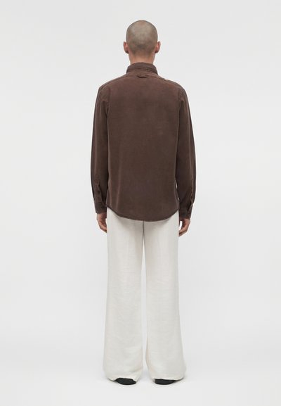 Άνδρας με ξυρισμένο κεφάλι που φοράει ένα μακρυμάνικο καφέ πουκάμισο και άσπρο παντελόνι με φαρδιά πόδια, στέκεται με την πλάτη προς τα έξω σε απλό φόντο.