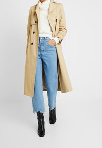 Beige trench coat with double-breasted buttons, paired with a cream knit sweater and light blue high-waisted jeans. Black ankle boots.