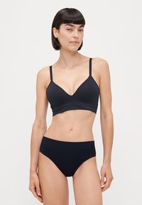 Melnā bralette ar plāniem siksniņām un V veida kakla izgriezumu, kopā ar atbilstošām augstām bikini biksēm, abas izgatavotas no gludas auduma.