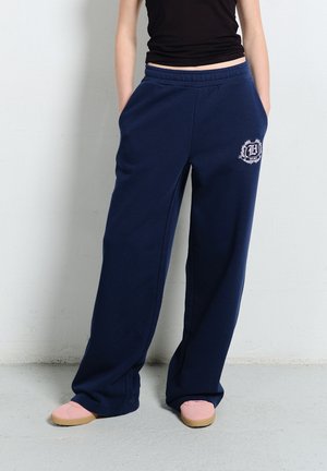 Osoba ubrana w granatowe, szerokie spodnie dresowe z logo na lewej uda, czarną bluzkę i różowe buty, stojąca przy białej ścianie.