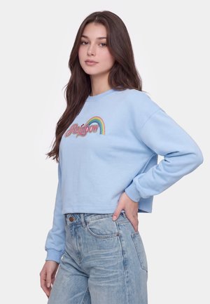 Ung kvinde med langt brunt hår iført lyseblå sweatshirt med "Rainbow" og en regnbuegrafik, kombineret med lyseblå jeans, stående med hånden på hoften.