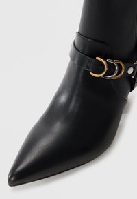 Bottes noires en cuir à la cheville avec un bout pointu et un talon fin. Présente une sangle décorative avec des accents en matériel doré et argenté.