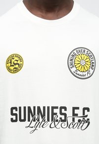 Bílé bavlněné tričko s černým nápisem "SUNNIES F.C. Lyle & Scott" a dvěma kruhovými logy s žlutými akcenty a květinovým motivem.