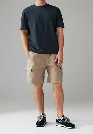 T-shirt en coton bleu foncé, short cargo beige avec poches latérales, et chaussures de sport noires avec des accents gris. Tenue décontractée présentée.