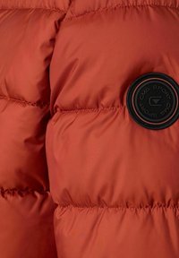 Detalje af orange dunjakke med en glat, skinnende finish, vandrette syninger og et rundt sort logopatch på venstre side.